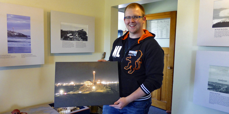 Braedon MacDonald at lighthouse, showing a photograph of the Dumping Day sendoff on the last Monday of November, when lobster fishermen drop their traps at sea.